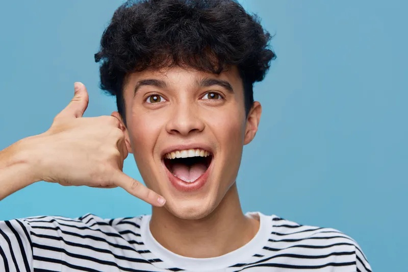 young man making call me gesture with hand near ear smiling wide against blue background, casual striped shirt, cheerful mood, communication sign