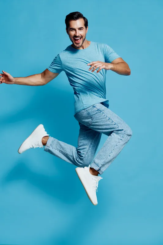 Happy cheerful young man jumping joyfully in casual blue t-shirt and denim pants on a bright blue background, expressing excitement and energy.