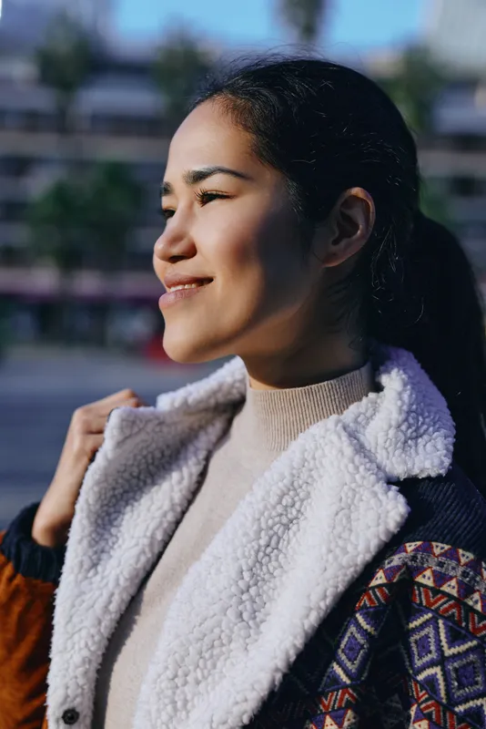 Smiling woman outdoors in a shearling jacket and knit sweater, basking in sunlight on a breezy day, showcasing casual fashion and relaxed confidence
