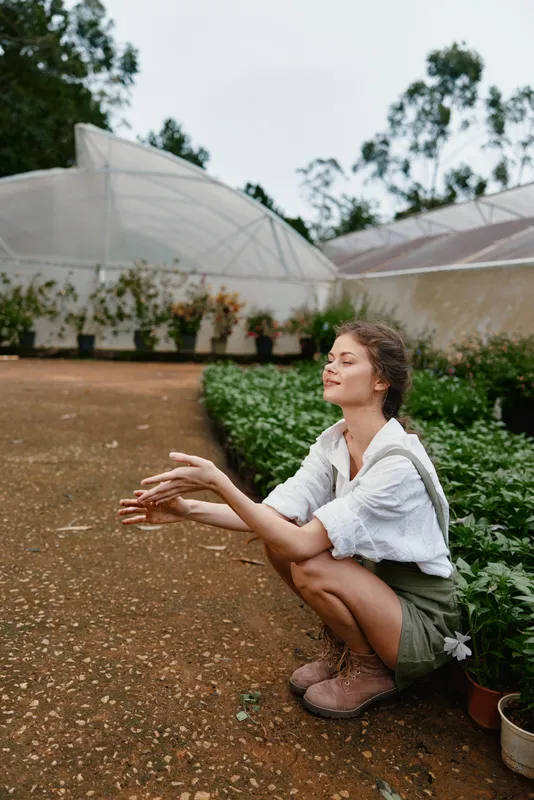 Young woman practicing mindfulness in a lush garden, wearing earthy tones and displaying a serene expression, surrounded by greenery and greenhouses