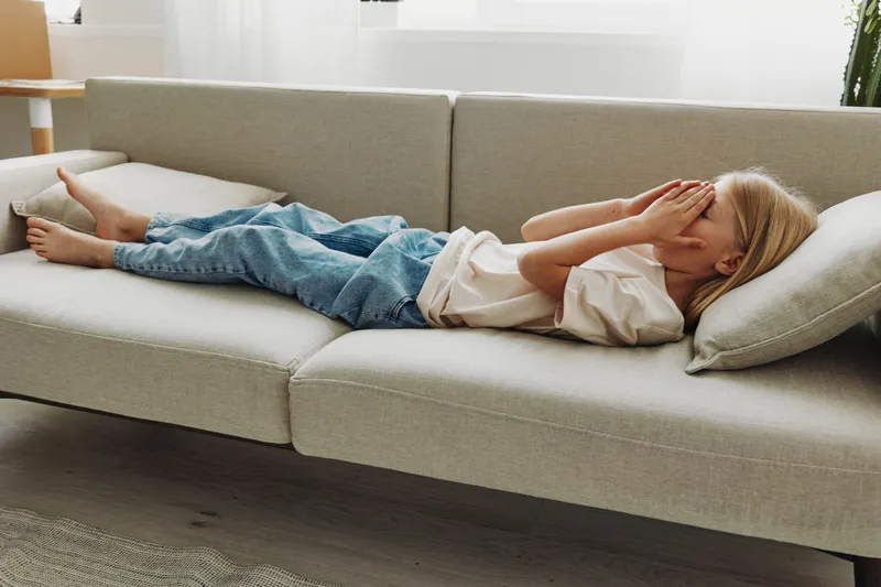 Tired child lying on modern sofa covering face with hands, wearing casual clothes in bright living room interior.