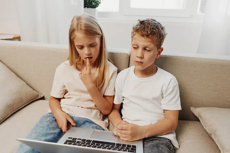 Curious children, boy and girl, sitting on a sofa using a laptop together in a bright room. Concept of learning, technology, and childhood exploration.