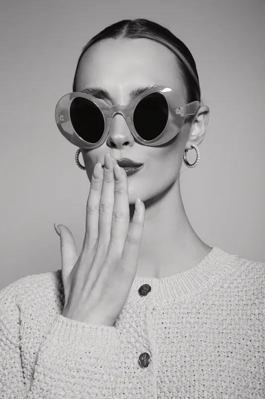 Young white woman with pale skin wears round sunglasses and knitted sweater. She covers her mouth with her hand in a stylish black and white portrait. Concept of fashion and mystery.