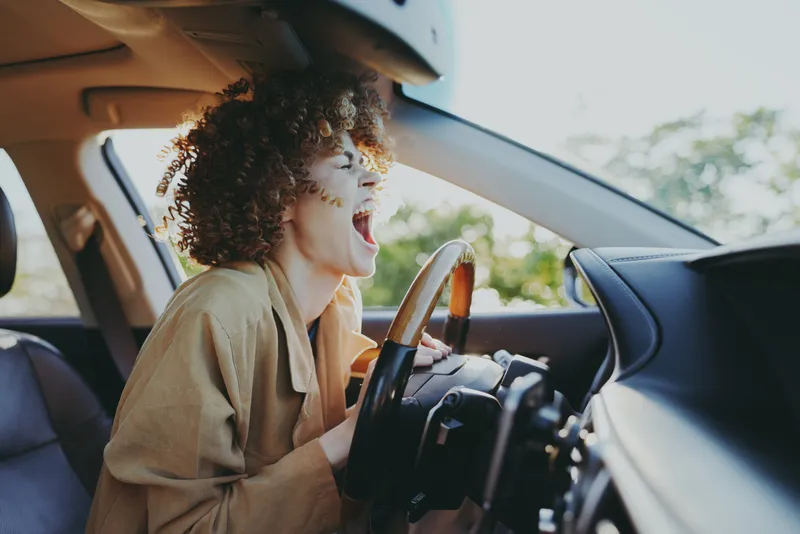 Bright candid scene of a woman driving, smiling with eyes closed, sunlit car interior, warm retro film color, casual outfit, relaxed mood, everyday adventure, natural light