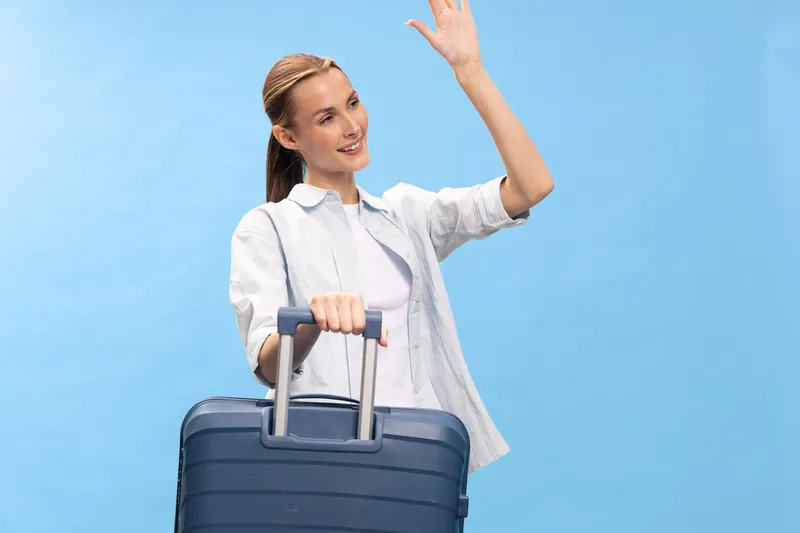 Happy young woman in casual clothes holds suitcase handle and waves hand, isolated on blue background. Concept of travel, vacation, tourism, and departure.
