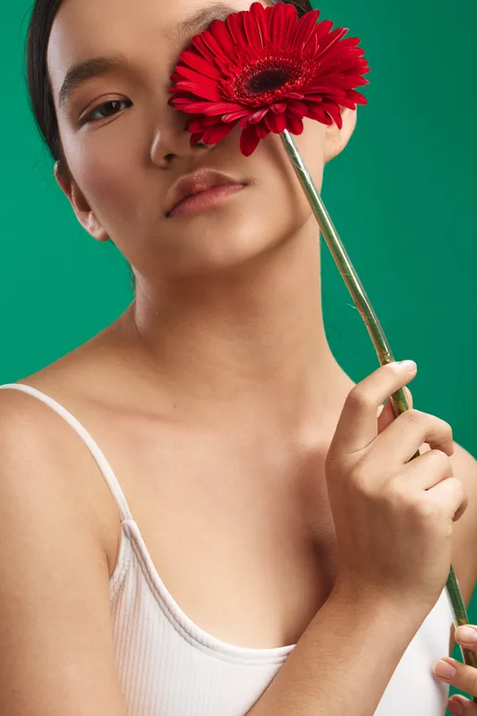 Asian woman delicately holds a red flower near her face, posing in a studio with a vibrant green backdrop to convey beauty, elegance, and a soft, expressive mood.