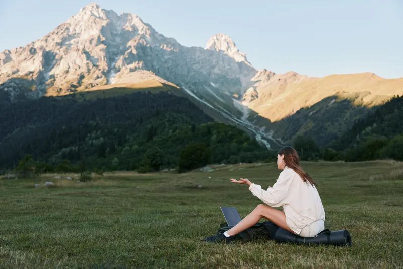 A relaxed traveler sits on a grassy meadow with a laptop, gazing toward distant mountains. The scene conveys focus, solitude, and outdoor inspiration in a tranquil setting.