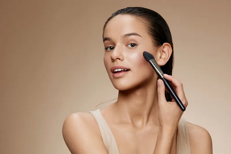 Young woman applying makeup with a brush, showcasing a natural beauty look on a soft beige background, expressing confidence and joy