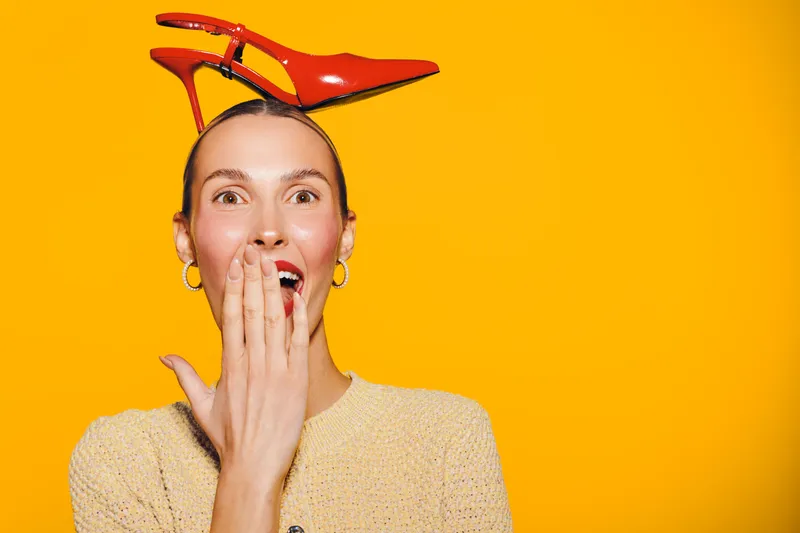 Surprised woman with red high heel shoe balanced on her head covers mouth with hand. Bright yellow background creates playful and quirky mood. Concept of fun, fashion, and unexpected moments.
