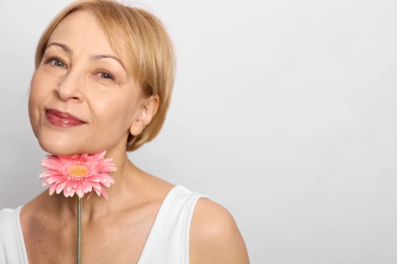 Cheerful blonde woman closeup portrait with a smile, holding a pink flower on a stick, soft light, beauty mood, clean white background