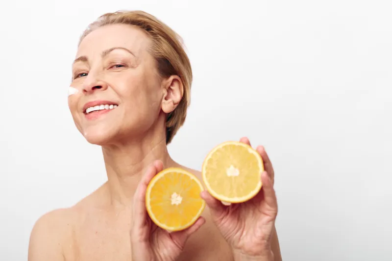 Smiling mature woman with short hair holds two orange halves close to face. Clean white background highlights fresh citrus fruit and natural skin care concept with healthy lifestyle and wellness.