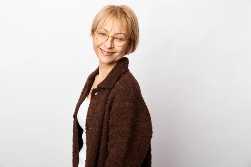 Woman portrait smiling glasses coat blonde mature fashion studio white background, female model in brown coat, relaxed friendly face, minimal studio lighting