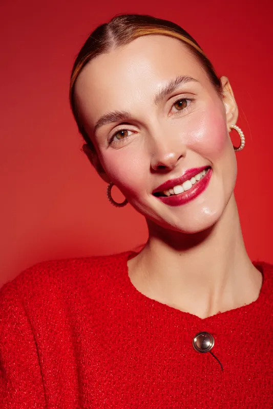 Smiling woman with clear skin and red lipstick wears red sweater and hoop earrings against red background. Portrait with natural makeup and confident expression in studio lighting.