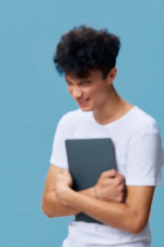 young man smiling and holding laptop close against light blue background, casual white t-shirt, curly hair, joyful expression in soft natural light