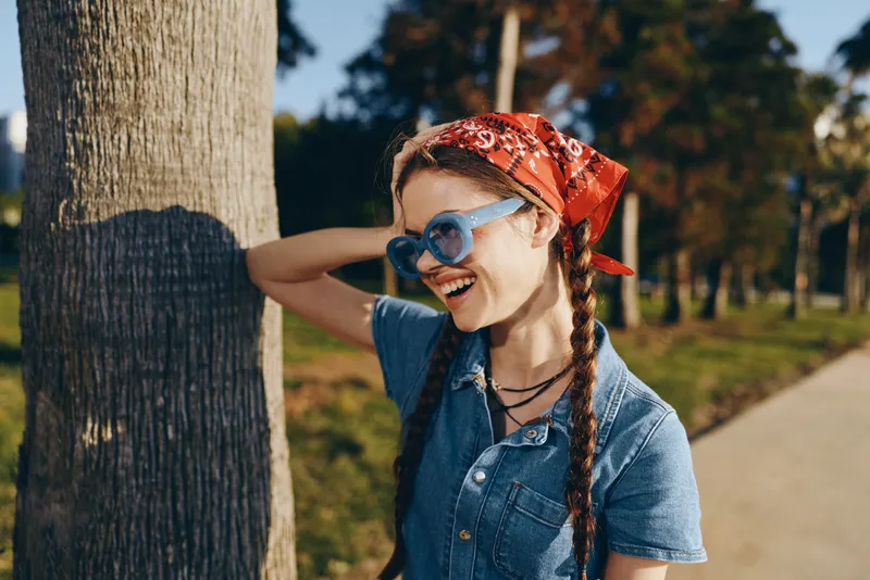 smiling woman with braids wearing sunglasses and red bandana standing by tree in sunny park, casual style, cheerful mood, natural outdoor setting