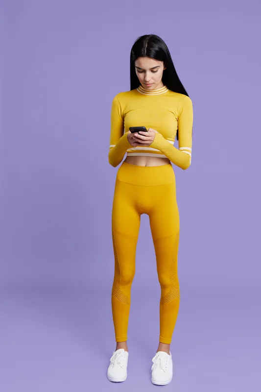 Sport fitness woman in yellow activewear standing against purple background looking at phone, showcasing modern workout outfit and slim silhouette, indoors studio setting.