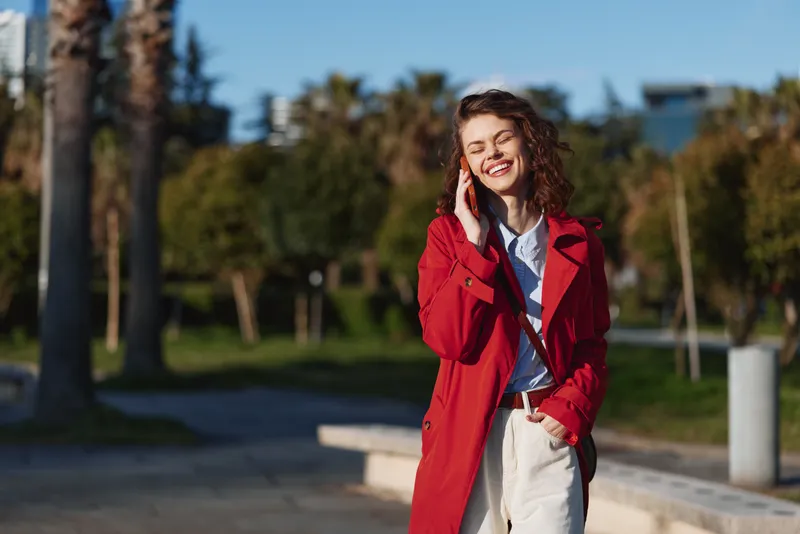 Happy woman outdoors walking in sunlight wearing red coat smiling with eyes closed enjoying fresh air urban park background casual style autumn day