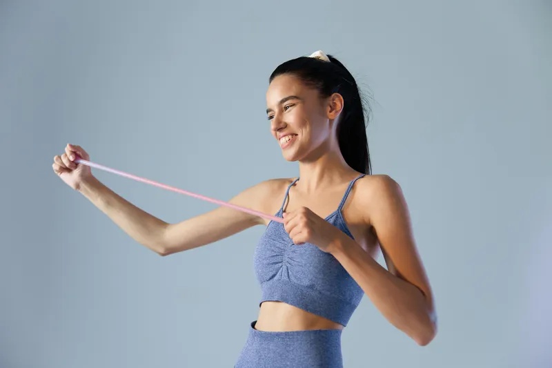 young woman sport fitness stretching with resistance band smiling and focused on light blue background, active healthy lifestyle concept