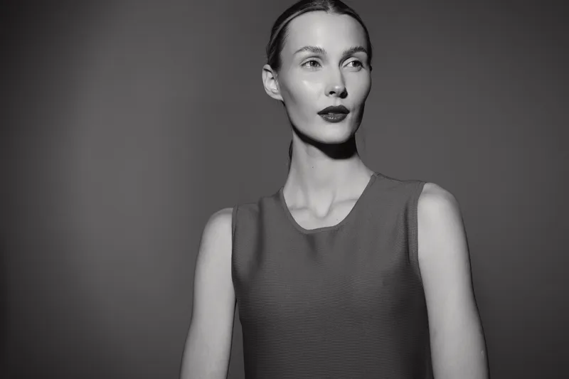 Tall slender white woman with pale skin poses in sleeveless top against dark background in studio lighting.