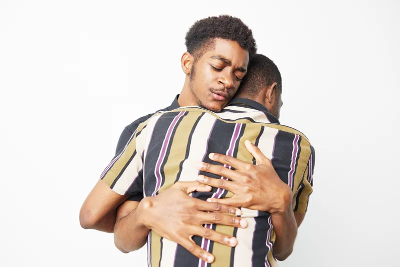 Tender embrace between two black men, conveying love and support. The vivid colors of their striped shirts contrast beautifully against the light background.