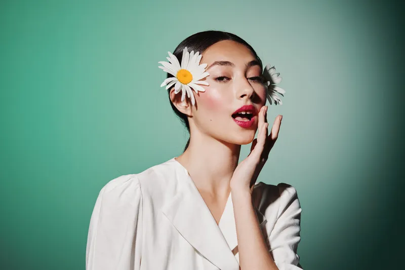 Young woman with bold red lipstick and white blouse poses with white daisy flowers in her hair. Studio portrait on green background in creative beauty fashion concept.
