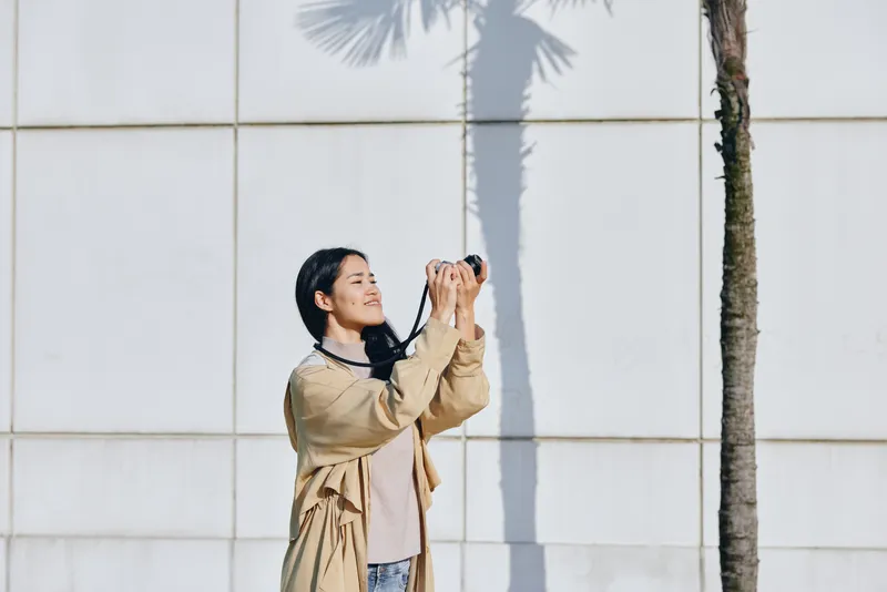 Camera, photography, outdoors, female in casual attire captures a moment under palm shadows beside a light wall, daylight glow, street vibe and candid motion