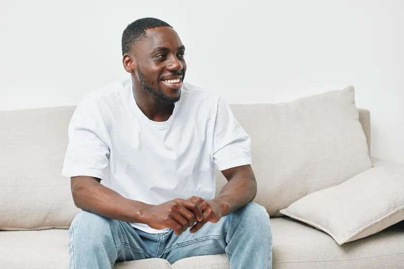 Smiling African American man in casual white t shirt and jeans sitting on a neutral colored sofa, expressing joy and relaxation at home