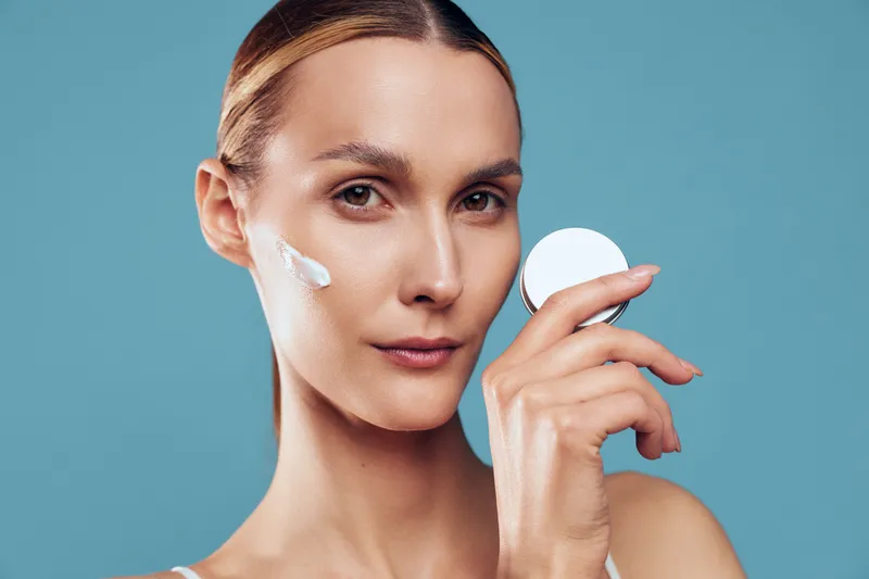 Beautiful young woman skincare routine, close up face portrait, applies white cream with cotton pad, clean hygiene concept, soft studio light on blue background