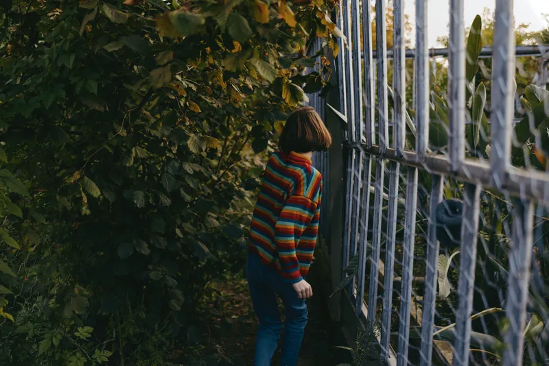 Child in colorful striped sweater with bob haircut leaning and peeking through metal fence amid dense foliage in a suburban backyard, curious expression, casual outfit exploration.