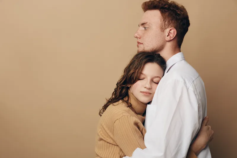 Young couple embraces tenderly against beige background. Woman with closed eyes leans on man wearing white shirt. Concept of love, comfort, trust, and emotional connection between partners.