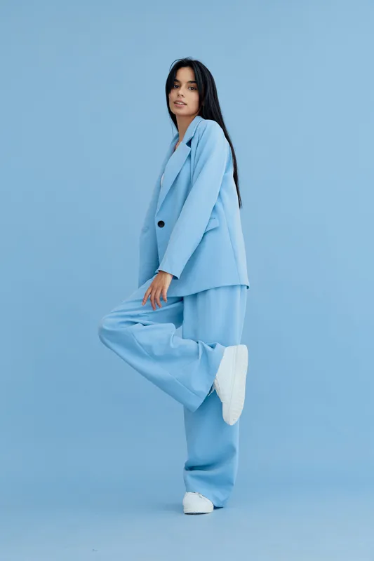 young woman in blue suit stands on one leg against blue background, wearing white sneakers and long straight hair, stylish casual outfit with modern minimalism