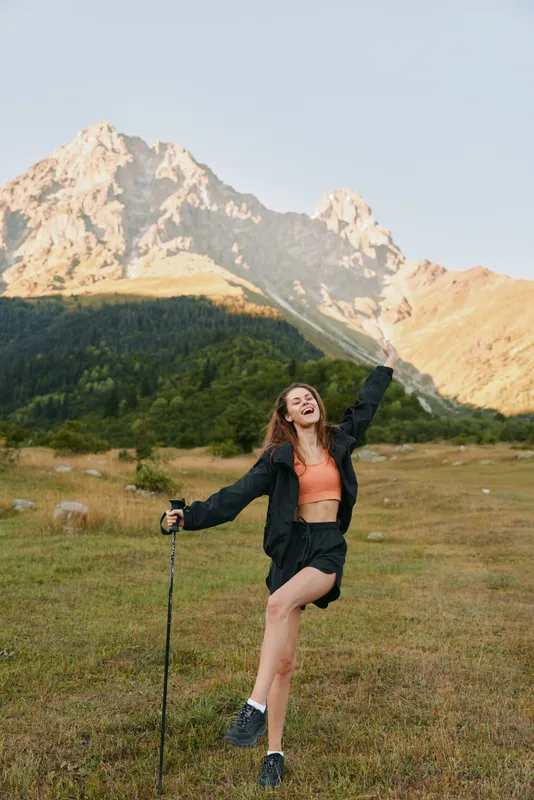 A joyful young woman hikes in a mountain landscape. She smiles and raises her arm in triumph beside a trekking pole, while rugged peaks rise behind a green field.