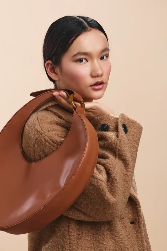 Asian woman wearing a chic wool coat poses with a round brown handbag against a neutral backdrop, exuding fashion elegance and a confident, modern vibe.