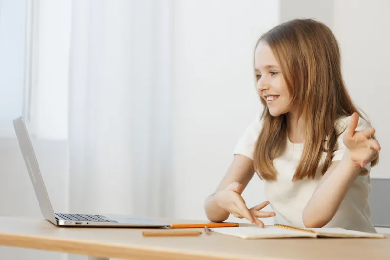 Happy girl engaging in a video call from home, with a laptop and notebook, showcasing a positive learning environment and creative expression