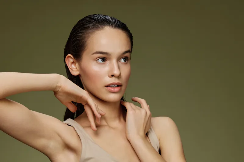 Young woman with fresh, glowing skin showcasing a natural beauty look, wearing a beige top against a soft green background, expressing confidence and serenity