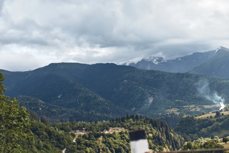 Serene mountain landscape with forested hills and distant peaks under an overcast sky, a small plume of smoke rising in a rural valley, vast natural scenery