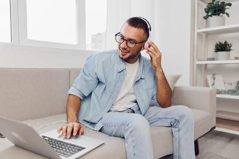 Engaged man working remotely at home, wearing headphones, with a laptop. Bright and cozy interior, showcasing a modern lifestyle and digital nomad vibes.