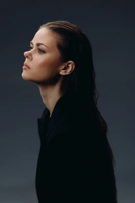 Profile of a serious woman with wet hair wearing a dark top against a soft gray background.