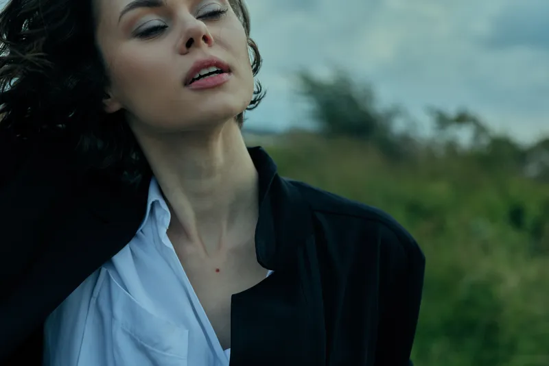 Woman in a black jacket and white shirt stands outdoors, eyes closed as wind moves her hair, moody natural light, fashion mood, solitary moment amid a field backdrop, calm and contemplative