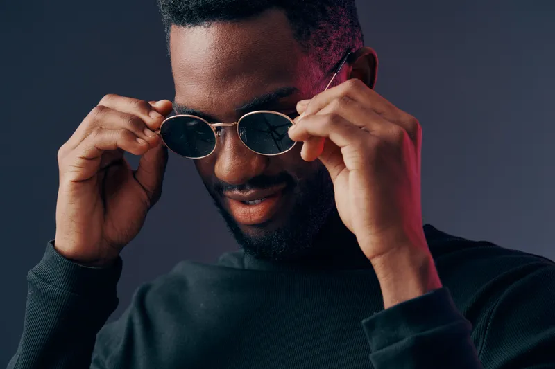 Stylish young Black man adjusting sunglasses with a confident expression against a dark background Perfect for fashion and lifestyle themes