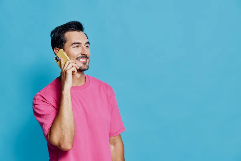 Smiling man wearing pink shirt talking on mobile phone, looking cheerful and happy against a bright blue background. Communication and lifestyle concept.