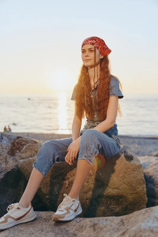 Lifestyle woman in modern boho-western denim street style sitting on rocks by the beach at sunset with film color effect and casual relaxed mood.