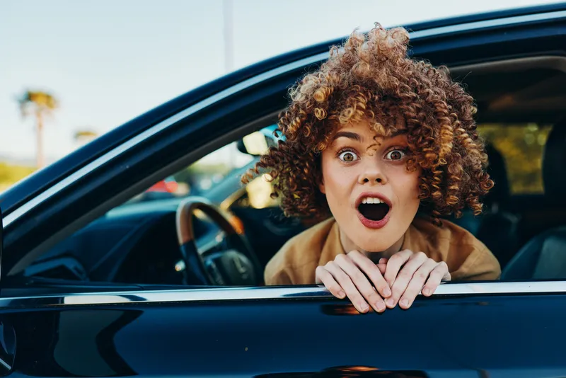 A young woman with curly hair leans from a car window, mouth open in surprise, captured in retro film color for a bold candid street moment.