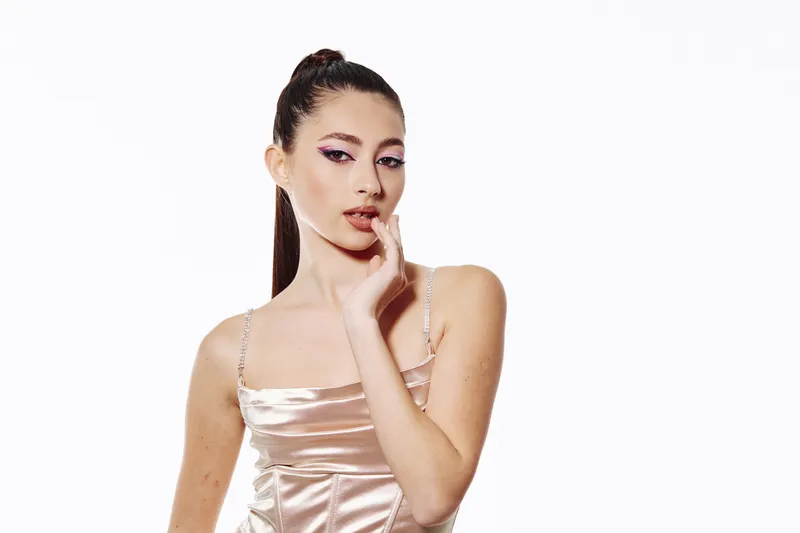Young woman with sleek ponytail and bold eye makeup in shiny satin dress poses with finger near lips on white background. Concept of beauty fashion and creative style.