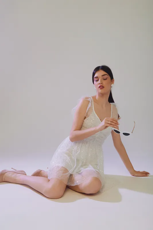 Young woman in a white lace dress poses on the floor holding sunglasses. Soft lighting highlights her beauty and fashion style on a neutral background.