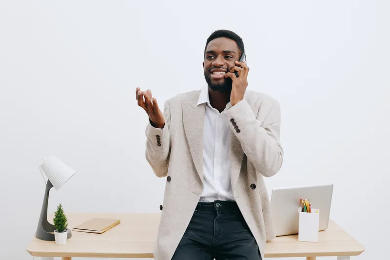 Confident smiling man talking on mobile phone while standing at modern office desk, casual beige blazer, white shirt, dark jeans, cheerful expression, bright minimalist workspace, professional lifesty