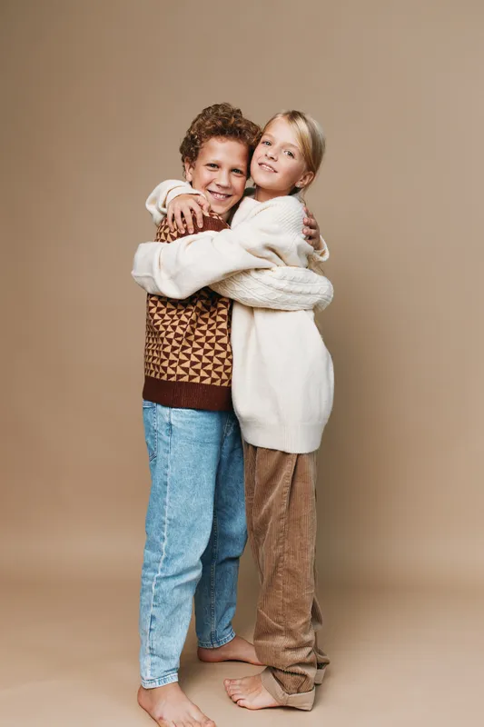Two children share a warm hug against a neutral backdrop, wearing cozy knit sweaters and casual denim, radiating comfort, friendship, and gentle affection.