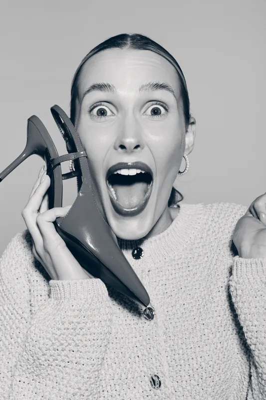 Emotional white woman with light skin holds shoe near face, mouth open in surprise, wearing knitted sweater on grey background.