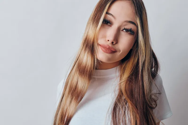Young woman with long hair and friendly smile wearing white shirt on light background. Closeup portrait of female model with natural makeup in simple studio setting.