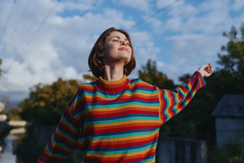 Young woman, short hair, striped sweater smiling with eyes closed outdoors, arms raised enjoying sunlight in suburban nature. Casual outfit, joyful lifestyle portrait for travel and leisure.